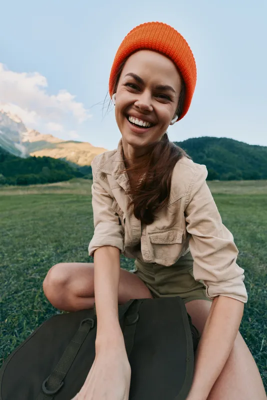 Cheerful woman wearing an orange beanie enjoys a sunny field landscape, presenting a casual lifestyle and an outdoor portrait with distant mountains in view on a clear day
