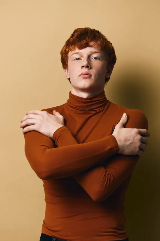 Young man with red hair wearing brown turtleneck posing with crossed arms against beige background. Serious expression, studio portrait with soft lighting and minimalistic style.
