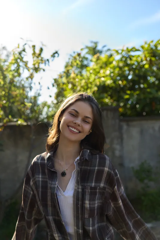 A cheerful young woman in a plaid shirt stands outdoors under sunlight, smiling warmly. A casual lifestyle portrait capturing happiness, confidence, and natural beauty in a bright setting