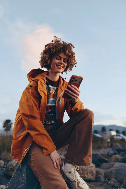 Woman lifestyle portrait outdoors, curly hair, orange jacket, smiling, holding smartphone, posing in rocky mountains under blue sky, with jeans and boots, autumn daylight