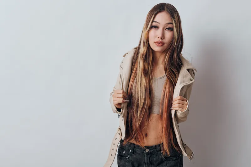 Young woman with long braided hair wearing a beige jacket and black pants standing against white wall, posing confidently with neutral expression and casual style.