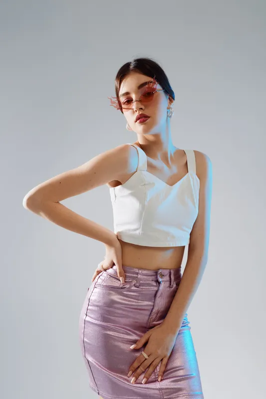 Young woman in stylish sunglasses and white crop top poses confidently wearing a shiny pink skirt. Studio shot on a neutral background with fashion and beauty concept.
