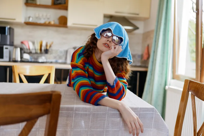 Girl in a colorful striped sweater with a blue towel on her head and sunglasses sits at a kitchen table indoors, conveying a playful, casual home lifestyle moment. Capturing a cozy, bright kitchen