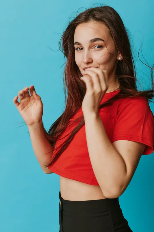 Young woman in red shirt posing with smile and playful gesture against blue background. Casual style, long hair, confident expression, studio portrait with natural light and relaxed mood.
