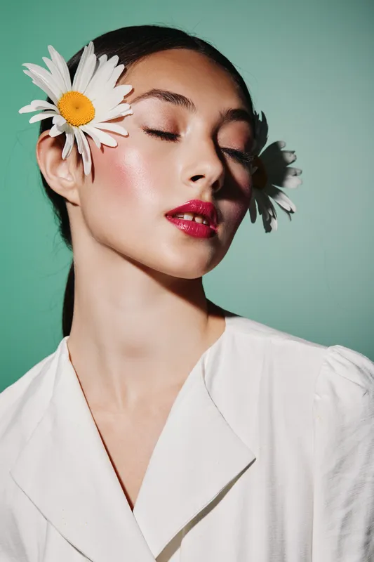 Young woman with daisies in her hair and vibrant makeup poses with closed eyes against a green background. Concept of beauty, fashion, and creative style in studio.