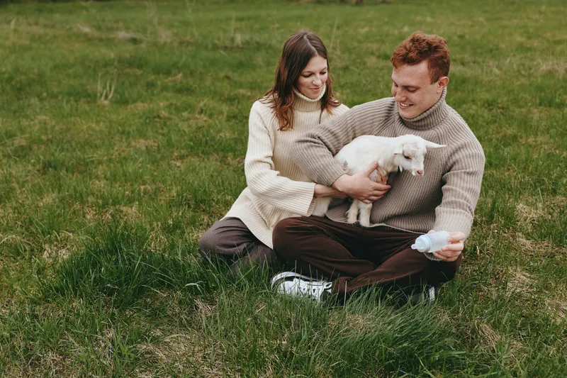 Happy couple outdoors on a sunny day, sitting on grass in a park, a small white puppy between them, wearing cozy sweaters, casual lifestyle, warm and affectionate moment