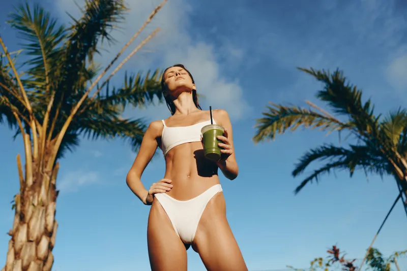A confident woman in white bikini stands outdoors under palm trees holding a green smoothie. The bright sunny day and clear blue sky highlight a healthy lifestyle concept.