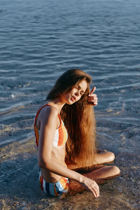 Woman on the beach by the ocean and sea, wet long hair draping her shoulder. She sits on the shoreline in sunlight, showing relaxation, serenity, summer wellbeing, and gentle self care.