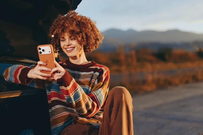 Woman lifestyle portrait of curly hair woman smiling with smartphone beside car outdoors, mountains at sunset in background, striped sweater, joyful travel moment
