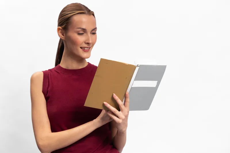 Smiling young woman in sleeveless burgundy top reads an open book with interest on white background. Concept of education, learning, knowledge, and personal development.