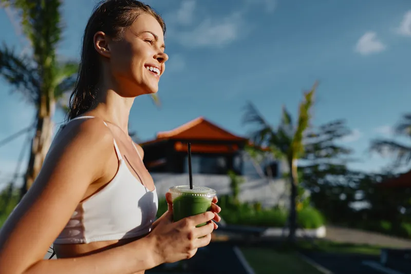 Cheerful woman outdoors, smiling with closed eyes, sunny moment, casual white sports top, holding a green smoothie, tropical backdrop with palm trees, bright colors, fresh lifestyle vibe.