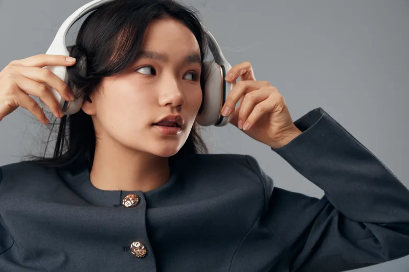 Young woman wearing headphones and dark blazer adjusting audio device with focused expression indoors against neutral background. Technology, music, sound and lifestyle concept.