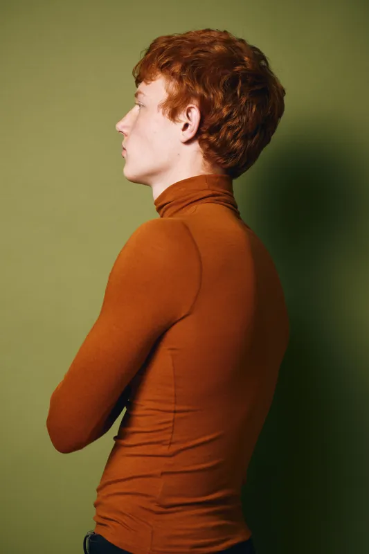 Young man with red hair wearing brown turtleneck standing in profile with crossed arms against green background. Side view, casual pose and contemplative expression.