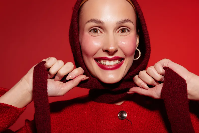 Smiling woman in red sweater and scarf poses on red background. Close-up portrait with bright lipstick and hoop earrings. Concept of winter fashion, happiness, and cozy style.