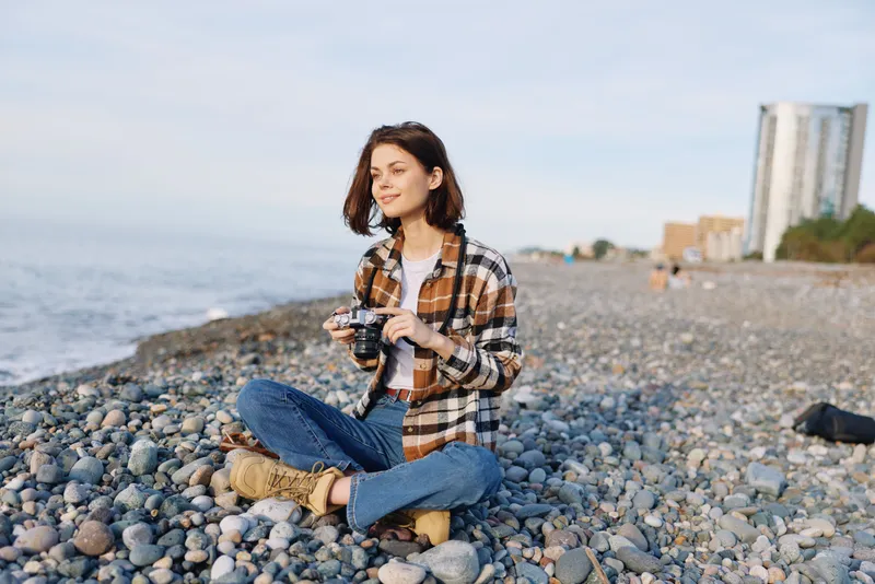 Woman photographer on a pebble beach with a camera, wearing a casual plaid shirt and jeans, sitting and enjoying the coastal view and calm sea breeze