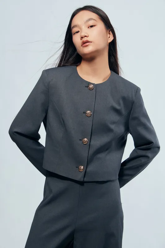Young woman posing in dark gray formal suit standing with hands behind back against light background. Professional style, modern fashion and confident expression for business or office look.