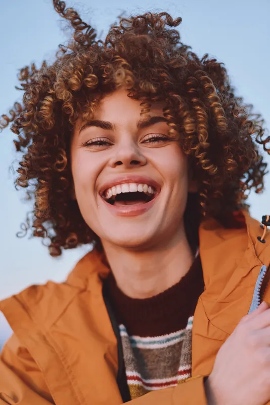 Woman with curly hair, smile outdoors, portrait of cheerful expression. Orange jacket and casual lifestyle convey happiness in natural daylight and sunny sky.