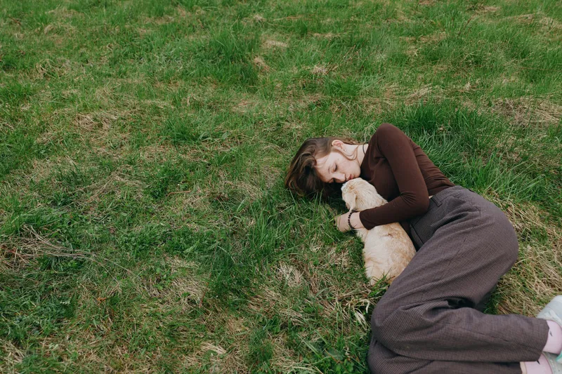 A relaxed woman rests on green grass, cuddling a small goat kid in a sunlit field. A candid lifestyle moment highlighting tenderness, care, and the bond between human and animal.