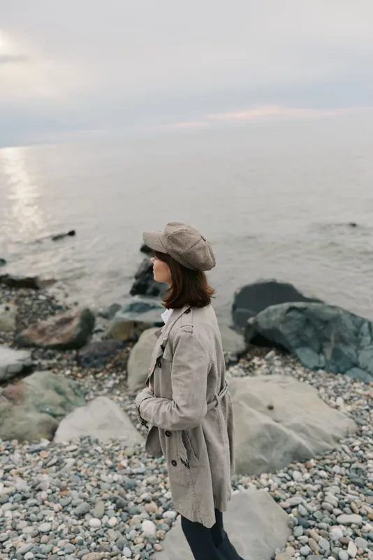 A woman in a beige coat and hat stands on a rocky beach looking out at the calm sea during cloudy weather. Concept of solitude, nature, and autumn fashion outdoors.