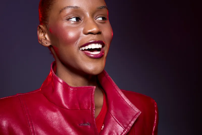 Smiling dark skinned African woman with short hair wearing red leather jacket looks away with joy. Studio portrait on dark red background. Concept of fashion, beauty, confidence, and elegance.