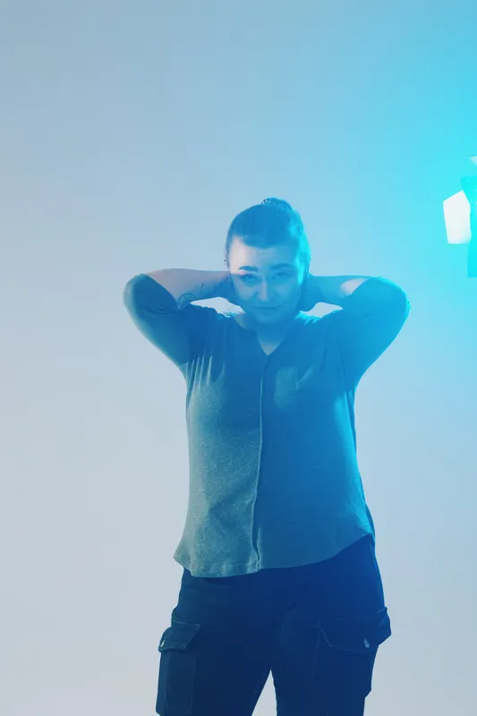 Young woman in casual fashion poses with hands behind head in a blue lit studio. The image highlights beauty and confidence with a modern style and soft lighting effects.