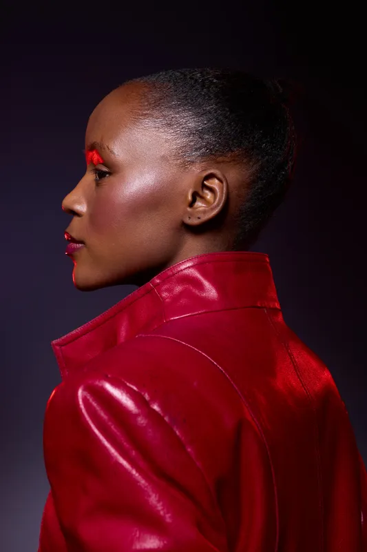 Profile of a dark skinned African woman wearing a shiny red leather jacket with bright red makeup on eyes and lips. Studio portrait with dramatic lighting and colorful fashion concept.