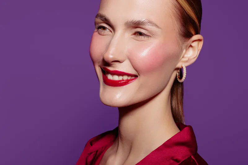 Smiling woman with glossy red lips and radiant skin poses against purple background. Close-up portrait highlights makeup, earrings, and elegant hairstyle. Concept of beauty and fashion.