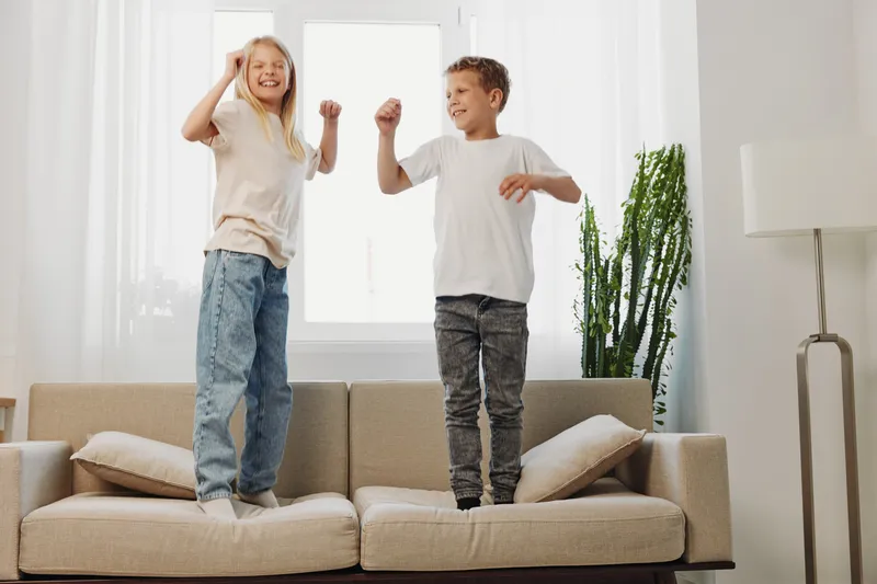 Happy children jump on sofa in bright living room, enjoying playful moment together near large window and indoor plant.