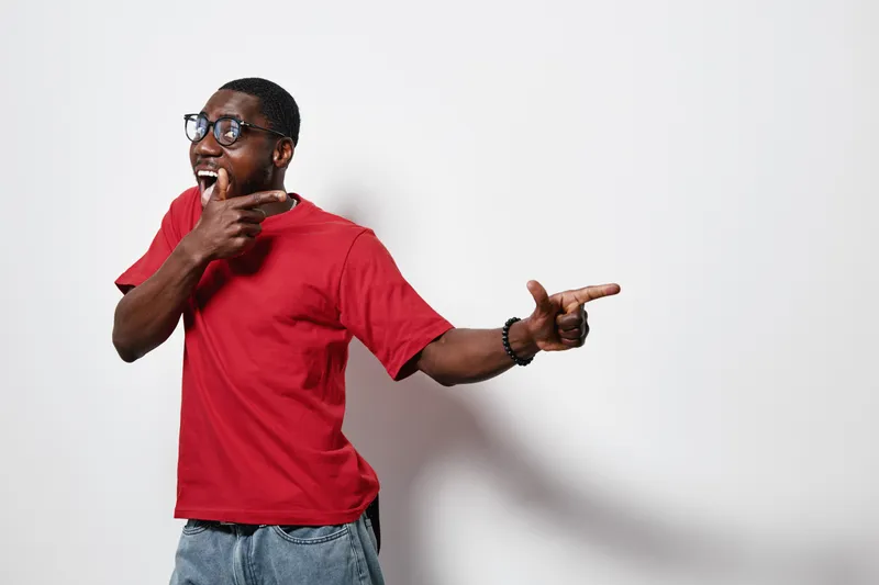 African American man with dark skin wearing red t shirt and glasses pointing sideways with excitement and smile on white background. Casual clothing and positive gesture.
