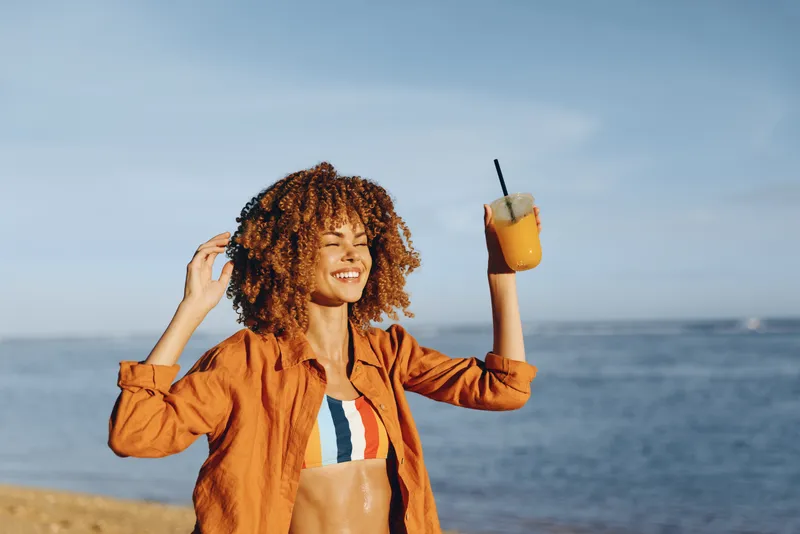 Smiling woman with curly hair wearing an orange shirt at the beach, holding a glass of juice with a straw. Sunny outdoor scene conveys carefree summer vibes and wellness and vitality.