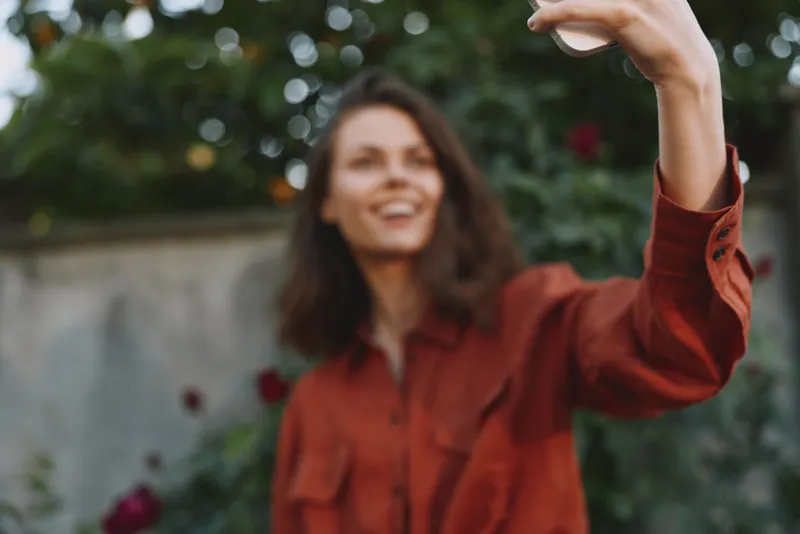 Cheerful woman outdoors, taking a selfie, wearing a red shirt, warm sunlight, joyful mood, relaxed pose, garden backdrop