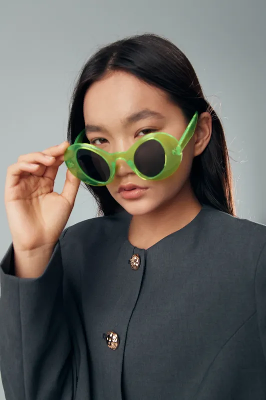 Young woman posing with green sunglasses and wearing a dark stylish jacket with buttons indoors, closeup portrait with neutral expression and straight black hair on grey background.