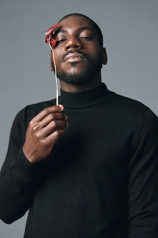 African man with closed eyes holding dried flower near face in creative conceptual visual style. Portrait of black male model wearing black sweater with calm expression on gray background.