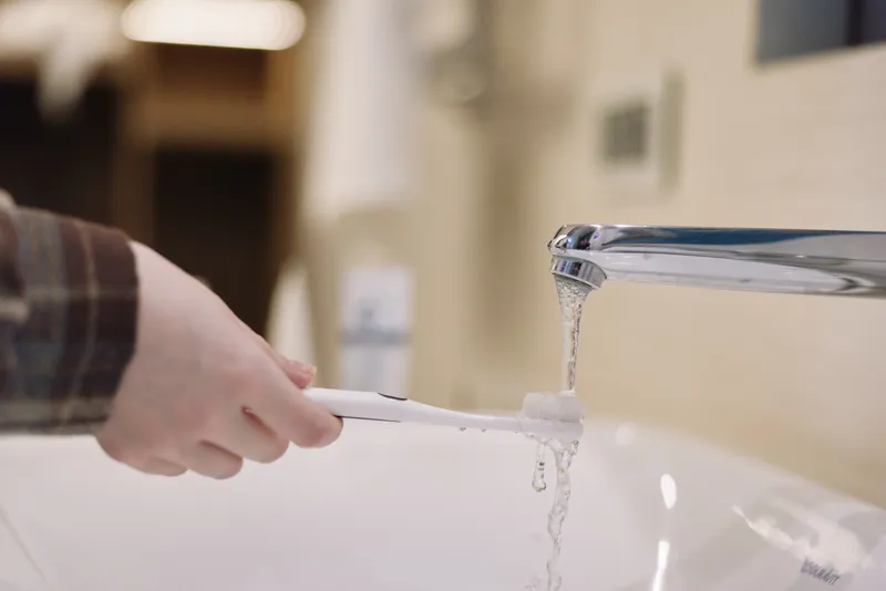 Hand holding a toothbrush under a running faucet, water pouring into a sink, focus on daily dental hygiene and routine care in a bright bathroom
