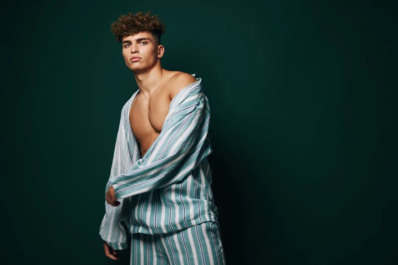 Young man with curly hair wearing striped pajamas posing against dark background, fashion portrait with casual style and confident expression in studio lighting.