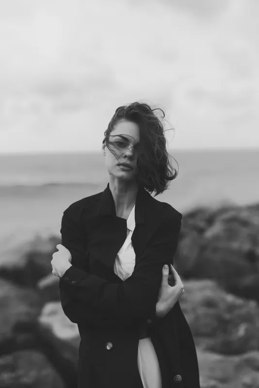 Black and white fashion scene with a solitary woman by the coast, trench coat over a white dress, windswept hair, moody seascape, minimalist mood, urban vibe meets nature, stylish and introspective.