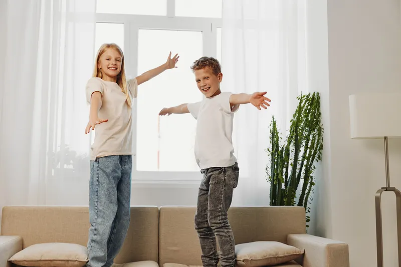 Happy children in casual clothes jumping on sofa in bright living room with natural light and green plants. Concept of joy, playfulness, childhood and family lifestyle at home.