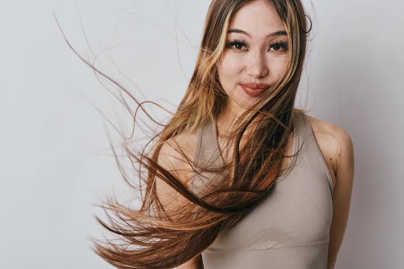 Young woman with long hair is posing with wind effect in studio. She is wearing sleeveless top and looking at camera with natural makeup and confident expression.