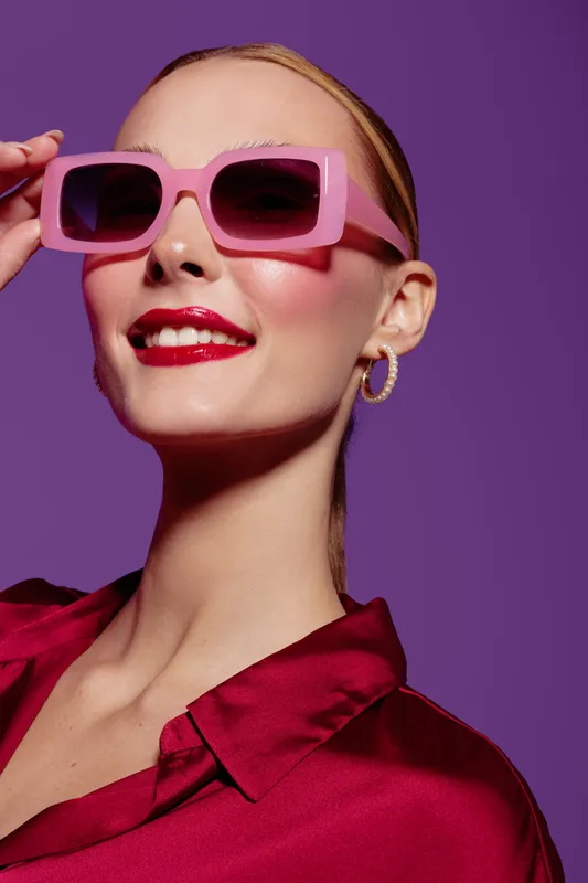 Stylish woman with pink sunglasses and red lipstick smiles confidently against purple background. Fashion portrait with bright makeup and trendy accessories in studio lighting.