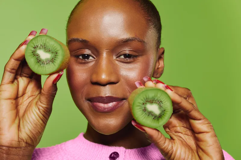 Close-up of smiling black woman holding two halves of fresh kiwi fruit near face. The healthy dark-skinned African female wears pink sweater against green background. Concept of wellness and natural