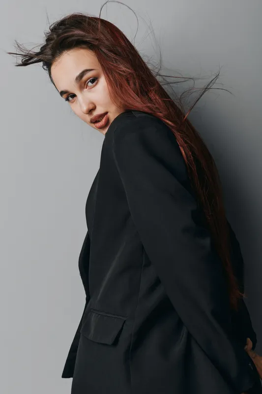Young woman with long hair wearing black blazer posing against gray background, looking over shoulder with confident expression and natural makeup in studio portrait.