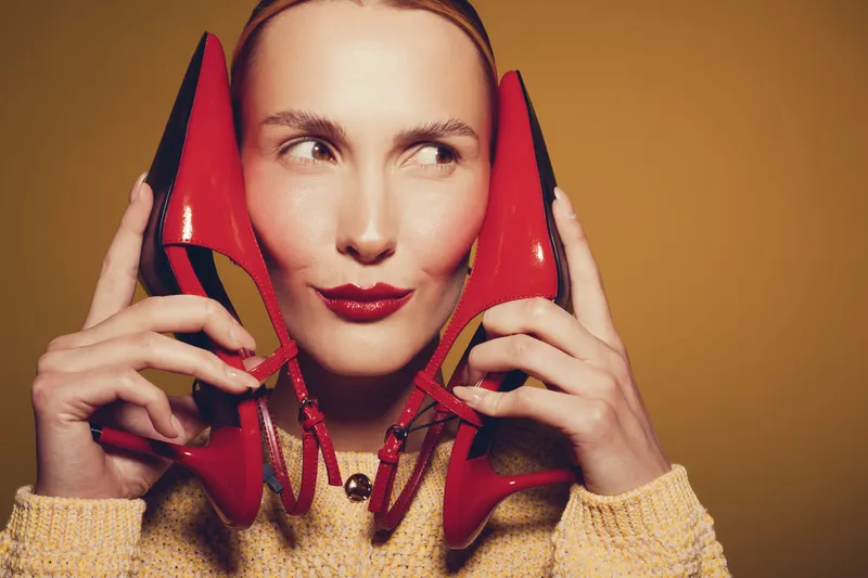 Young woman with red lipstick holds red high heels near her face, looking sideways. Concept of fashion, style, and playful mood with warm studio lighting.