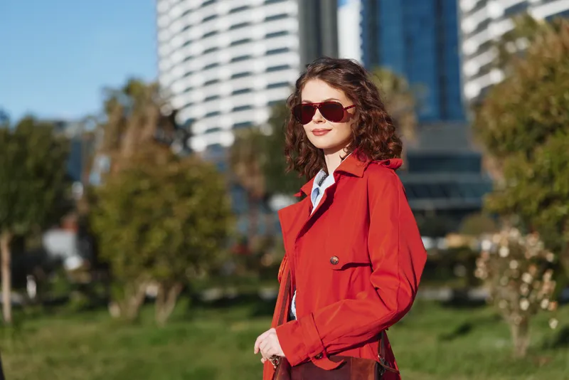 Woman in a red coat standing outdoors with sunglasses, urban cityscape behind. A confident, stylish portrait emphasizing fashion, street style, and sunny mood with soft natural light.