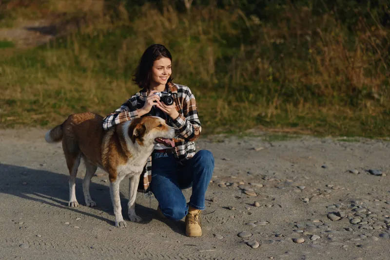 Woman kneels on a pebble beach, taking a photo with a camera while a large dog stands close, creating a warm moment of outdoor companionship and playful exploration.