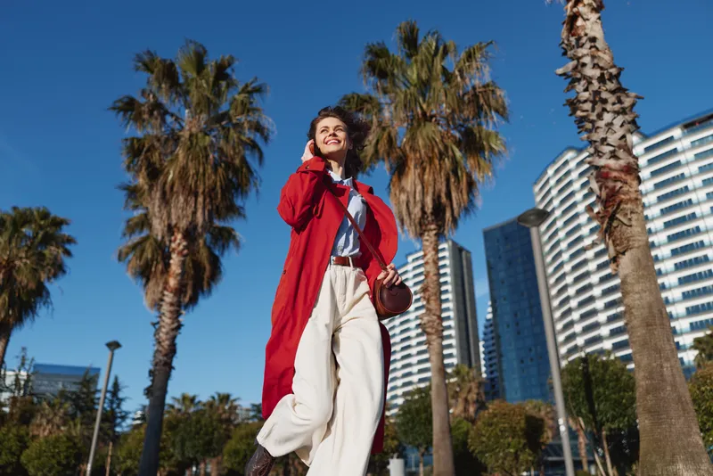 One woman in a red coat and white pants walks confidently through a sunny urban plaza with palm trees and tall buildings behind, her smile capturing city chic.