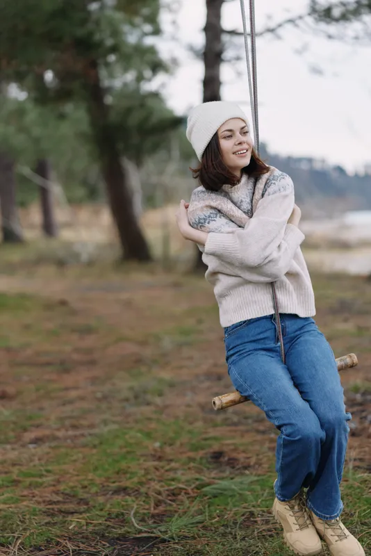 Woman on a swing in an outdoor forest setting wearing a cozy sweater and beanie, smiling and playful as she enjoys casual recreation among trees and pine needles