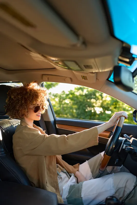 Casual woman drives a car in a sunlit interior, wearing a beige jacket and sunglasses, capturing a relaxed lifestyle with retro film color and candid urban vibe