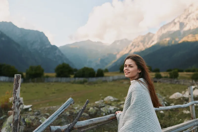 Young woman in a cozy gray cardigan stands beside a weathered wooden fence in a mountain valley, smiling softly as distant peaks glow under a clear blue sky, conveying a peaceful outdoor lifestyle.