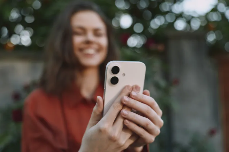 Cheerful woman outdoors, smiling while taking a selfie with a smartphone, soft natural light, casual outfit, relaxed pose, lush greenery background