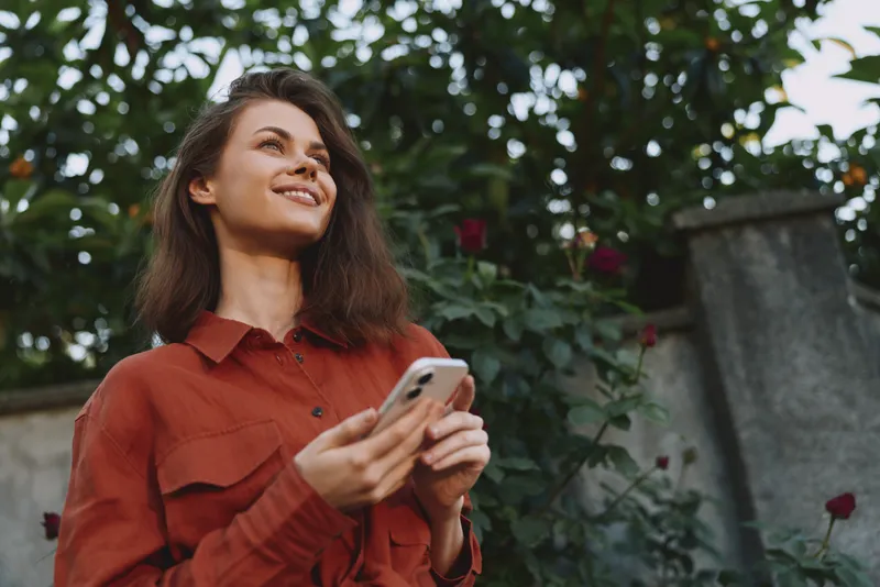 Young woman in a red shirt stands outdoors in a garden, smiling softly as she gazes up, holding a smartphone, enjoying a candid moment of connection with nature